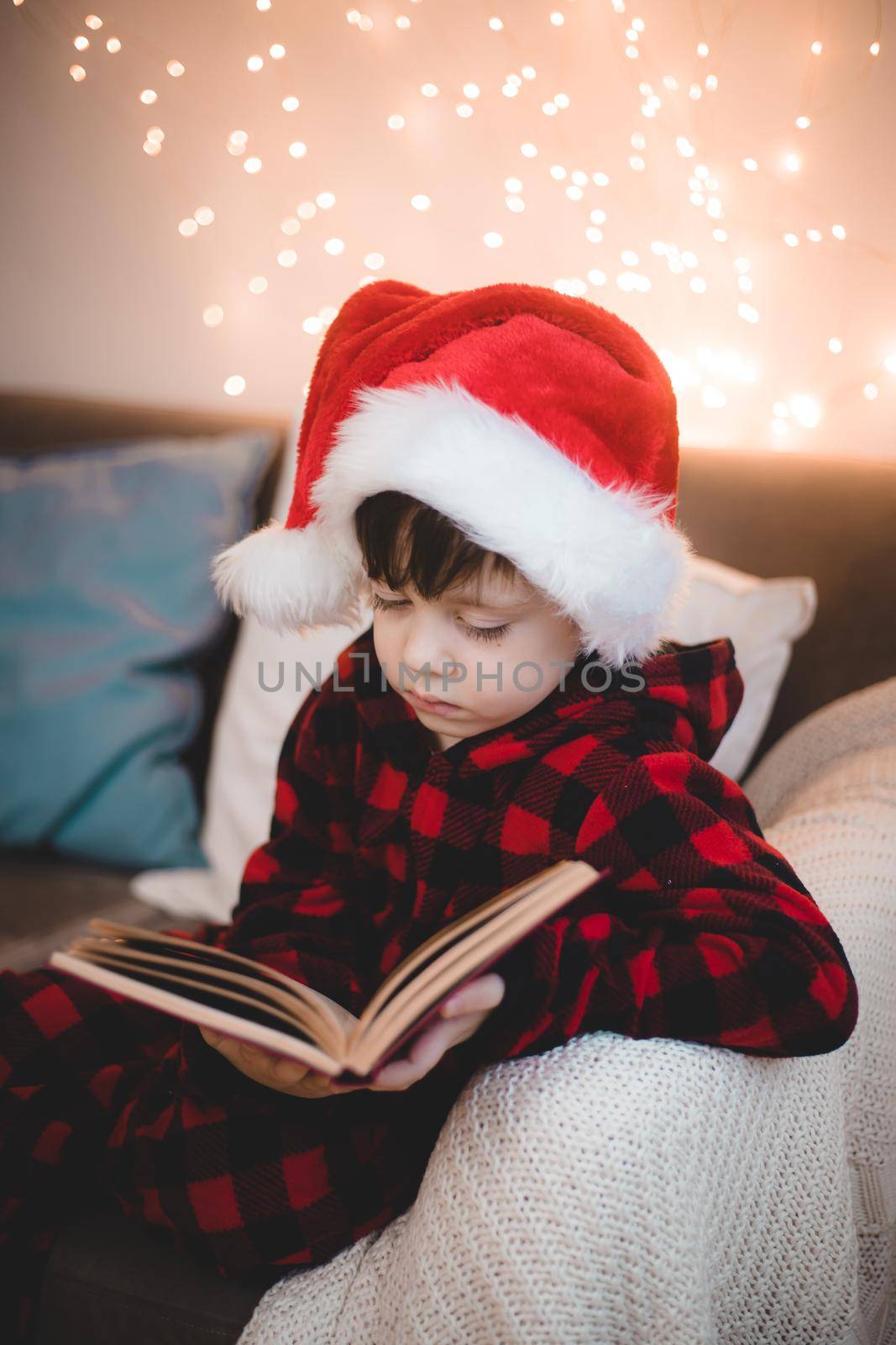 A boy in a Santa hat is reading a book on a lifestyle sofa. Reading books. New Year's mood. Garland on the wall