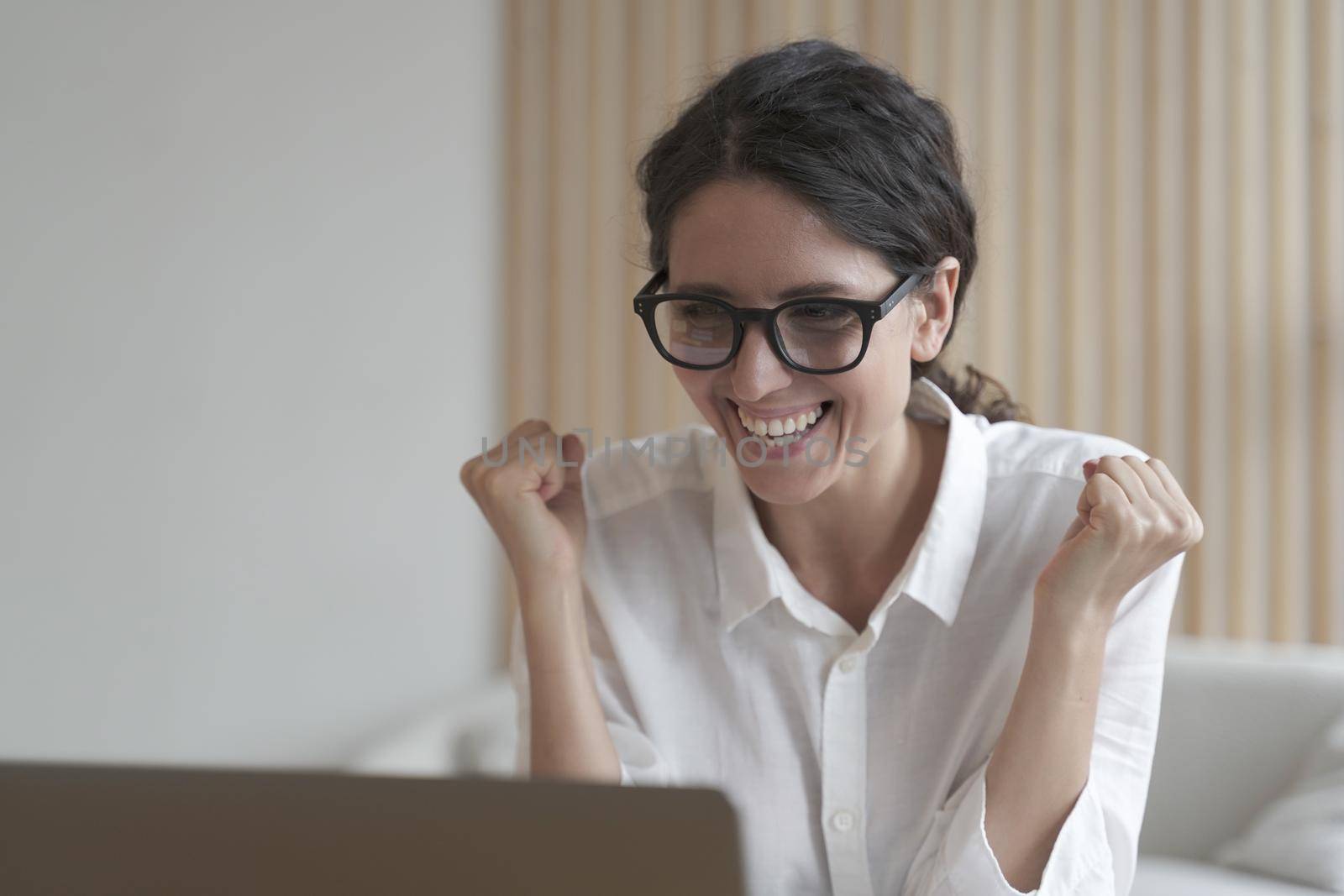 Excited italian woman in glasses looking at laptop screen and celebrating work success, receiving job offer by email, overjoyed female employee clenching fists while getting pleasant news on computer