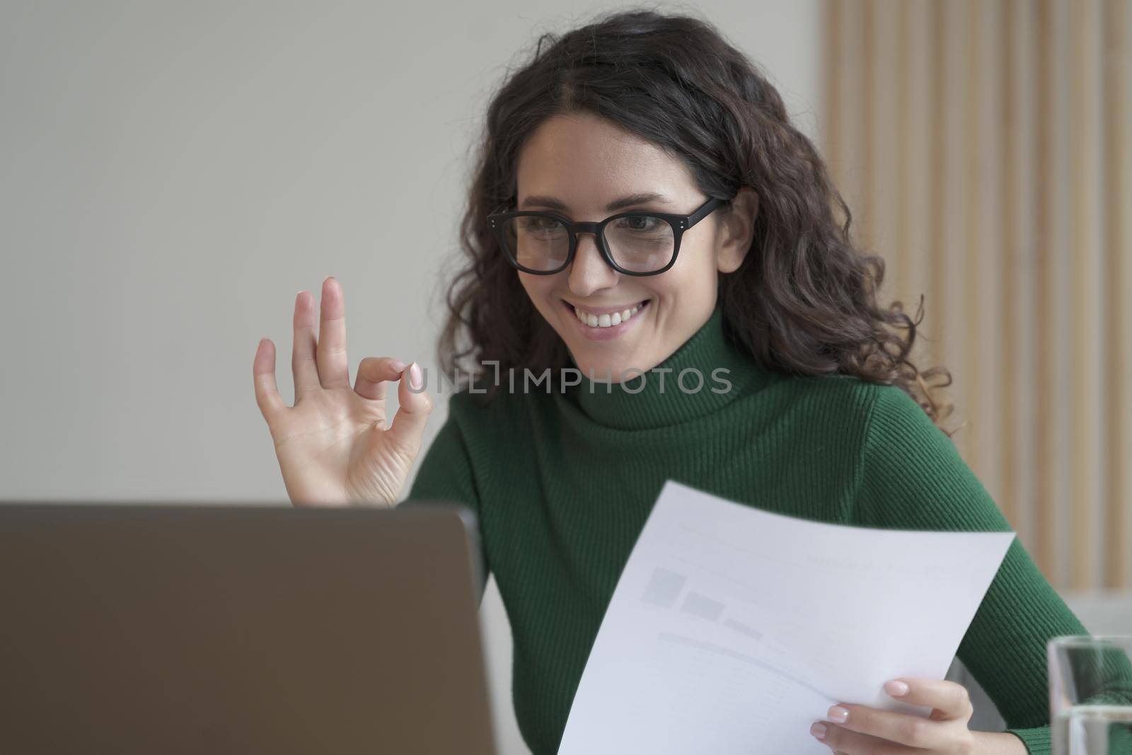 Smiling Italian female employee holding financial report and showing okay gesture during video call by vkstock