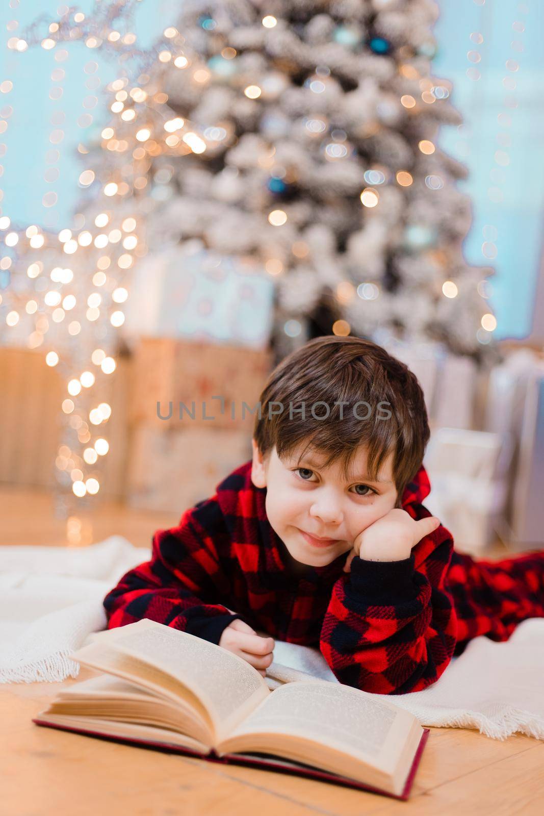A boy in pajamas reads a book under a Christmas tree . New Year's mood. Reading books . Children's books. by alenka2194