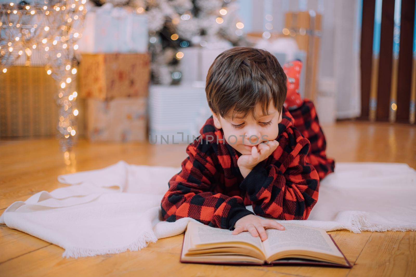 A boy in pajamas reads a book under a Christmas tree . New Year's mood. Reading books . Children's books. by alenka2194