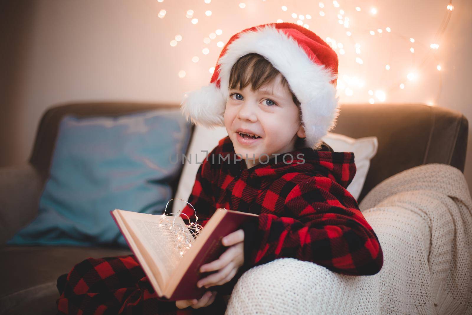 A boy in a Santa hat is reading a book on a lifestyle sofa. Reading books. New Year's mood. Garland on the wall