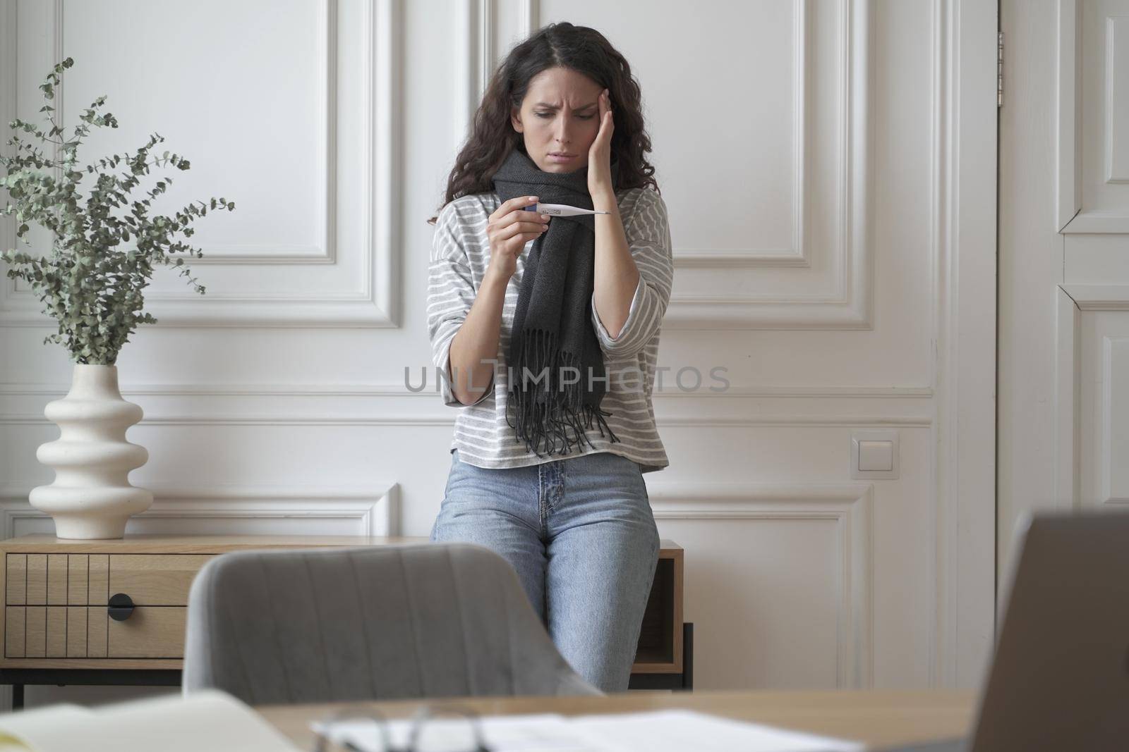 Unhealthy anxious business lady in warm scarf around her neck standing at home office worriedly looks at thermometer checking temperature touching forehead with hand, suffering from headache and flu