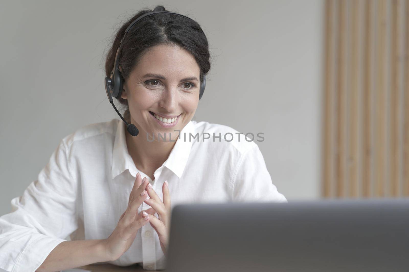 Smiling italian businesswoman in wireless headphones sitting at desk while works on laptop at home by vkstock