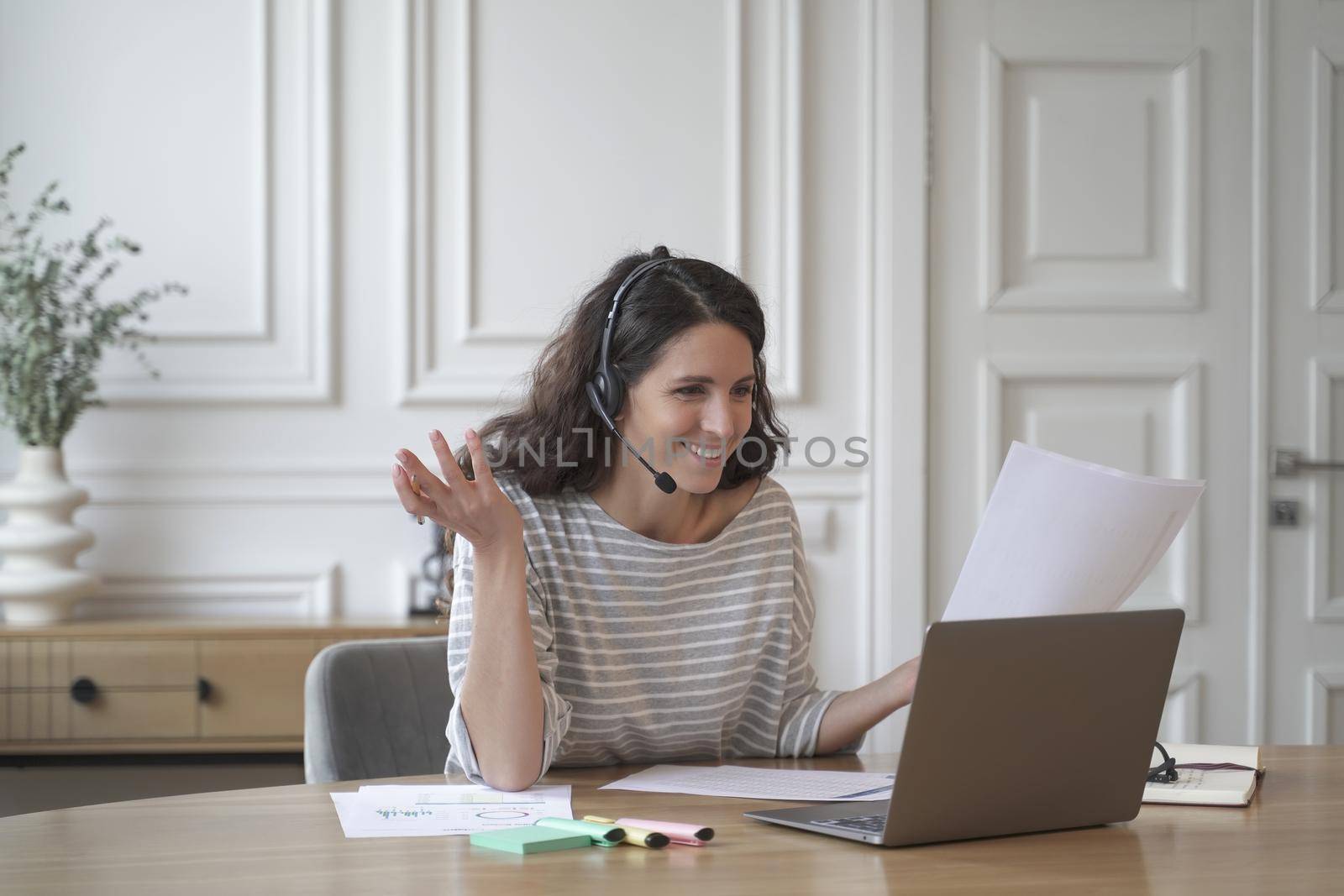 Young excited cheerful female couch in wireless headset with microphone conducts private consultations online on laptop while sitting at desk at home office. Freelance and distant education concept