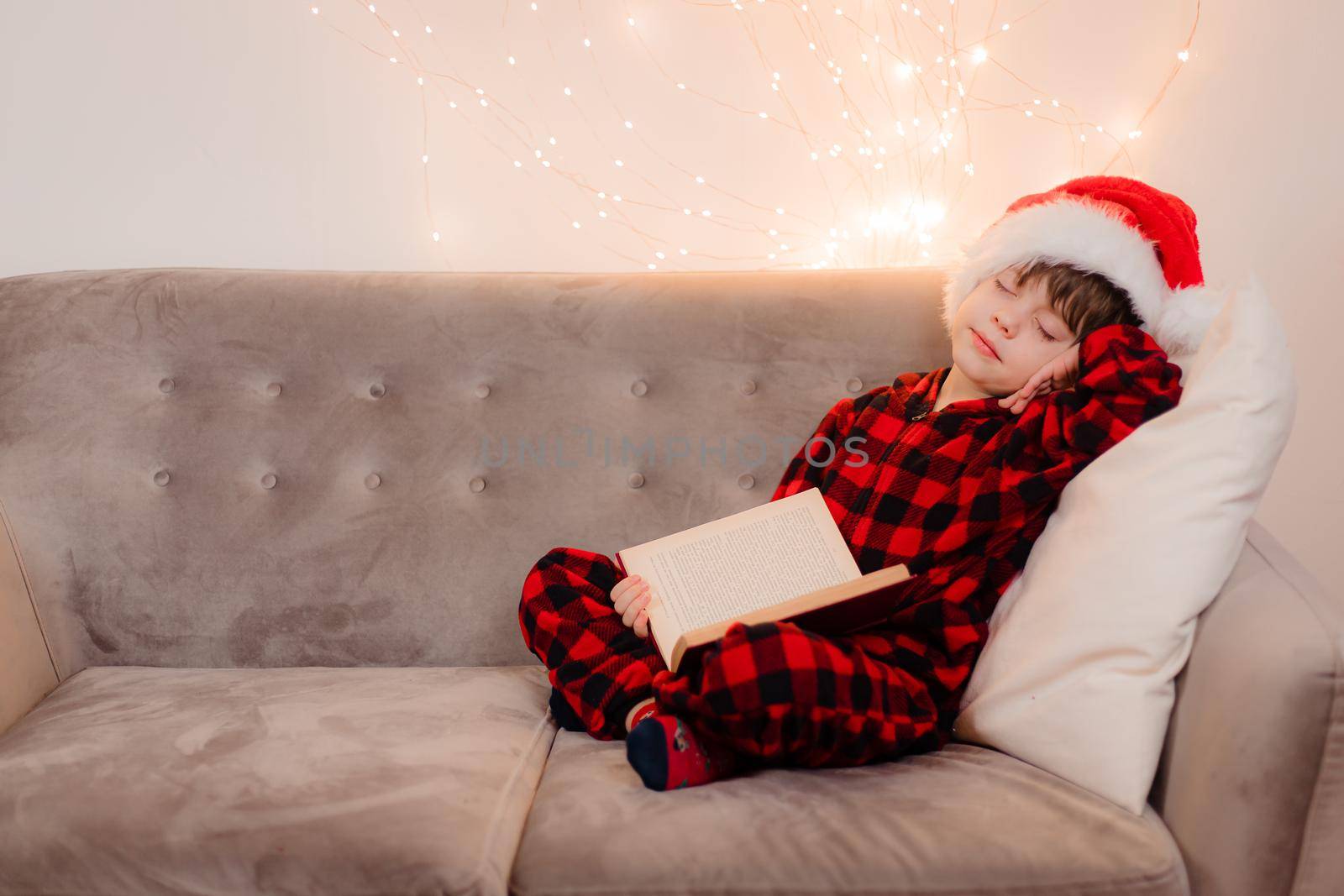 A boy in a Santa hat is reading a book on a lifestyle sofa. Reading books. New Year's mood. Garland on the wall