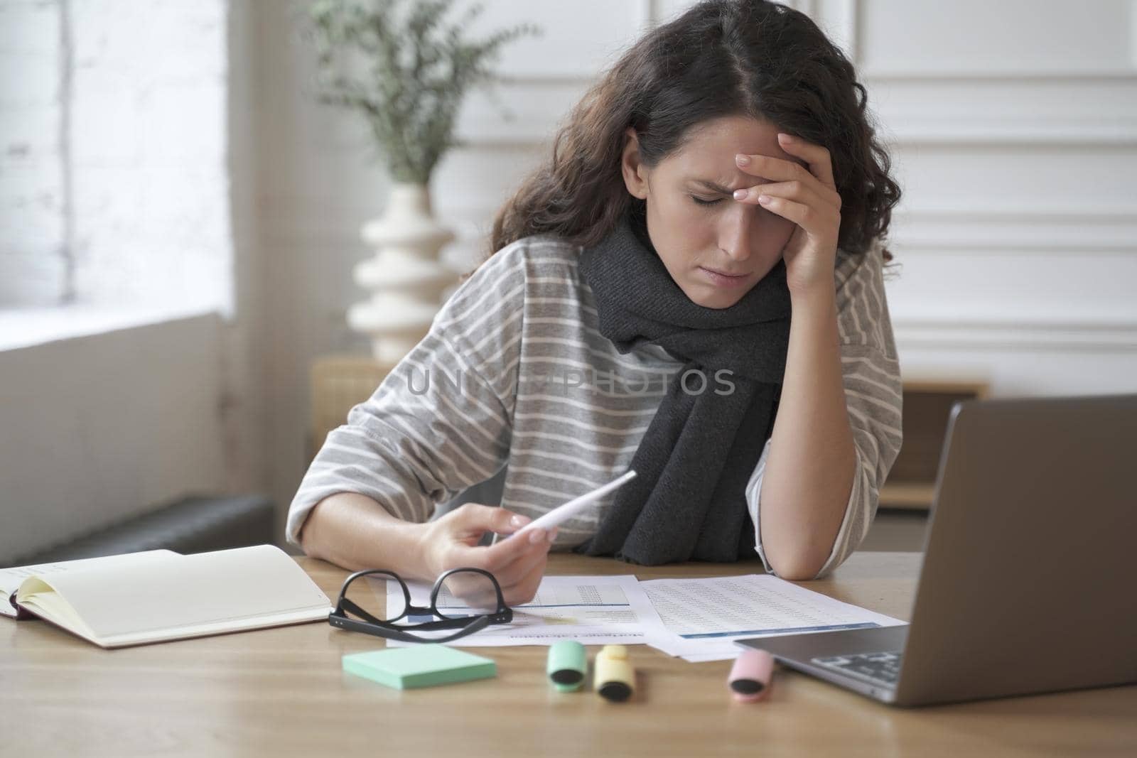 Young latin woman office worker sitting at table with digital thermometer in hand, suffering from headache and high fever at work, female emloyee feeling unwell sick, measuring temerature at workplace