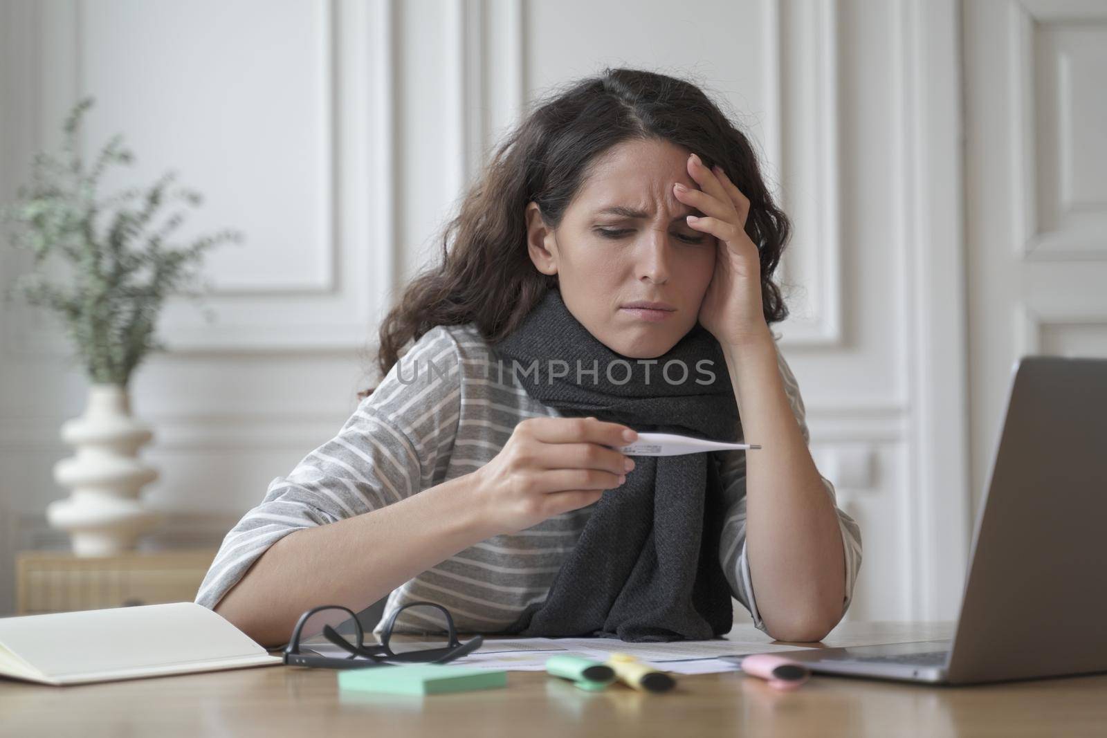 Unhealthy spanish woman wrapped in warm knitted scarf looking anxiously on digital thermometer by vkstock