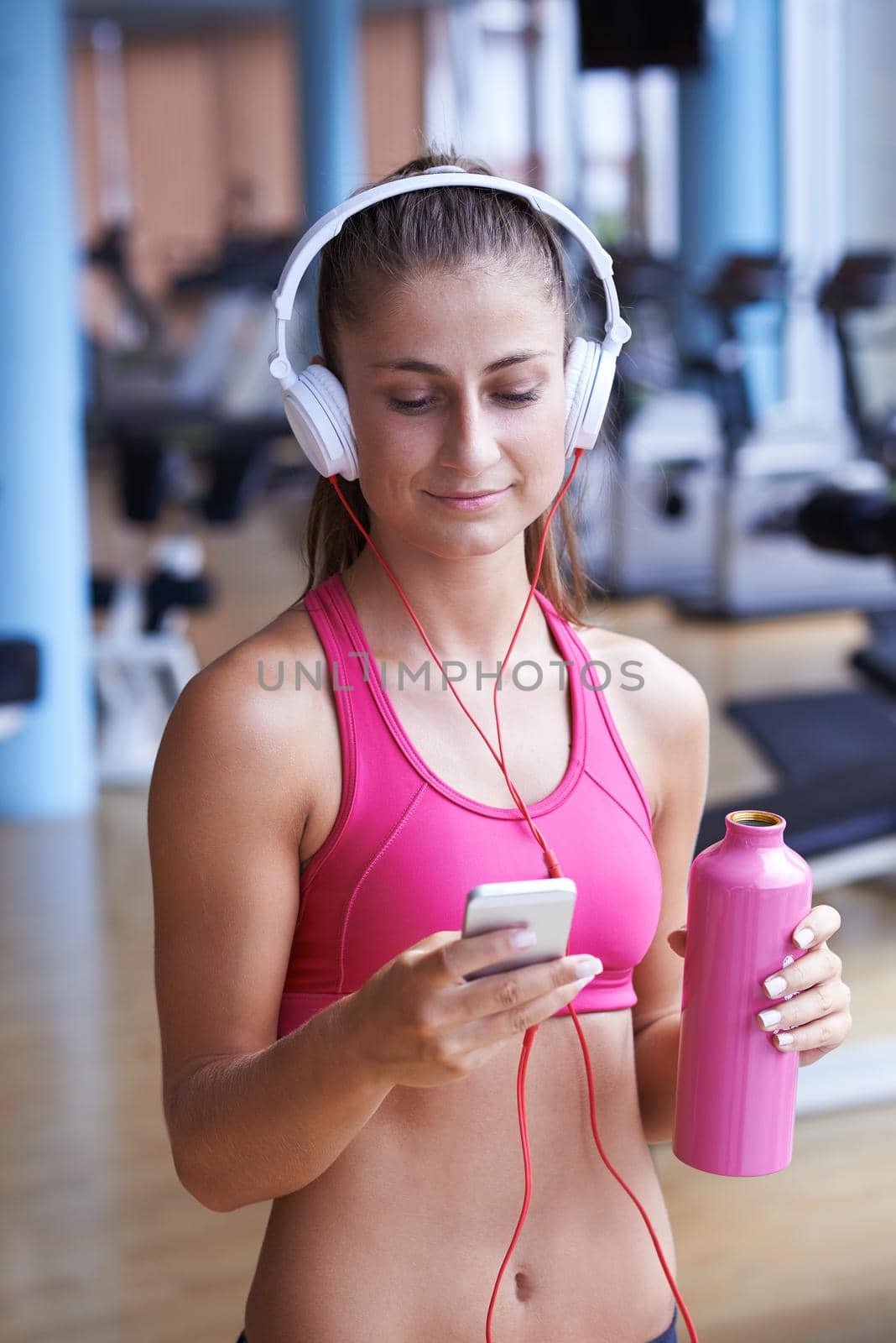 young healthy woman drinking water  in fitness gym while sitting on pilates ball and listening music on headphones from smartphone