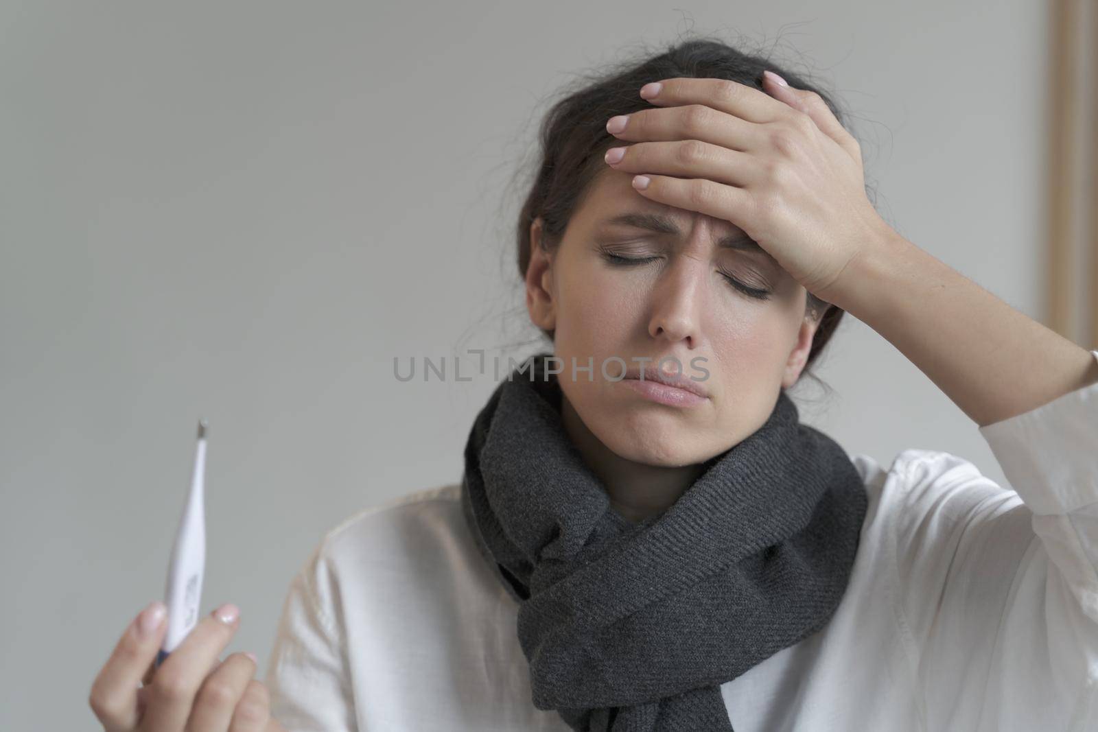 Italian entrepreneur lady tormented by hard work and fever, stands with closed eyes at home interior with warm scarf around her neck, while one hand on forehead and another holding digital thermometer