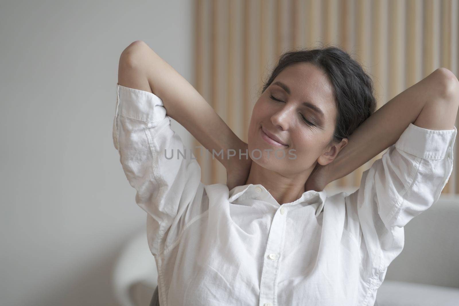 Close up of young happy Spanish woman with closed eyes dreaming, holding hands behind head, stretching body after hard working day in office. Smiling dreamy female resting at home