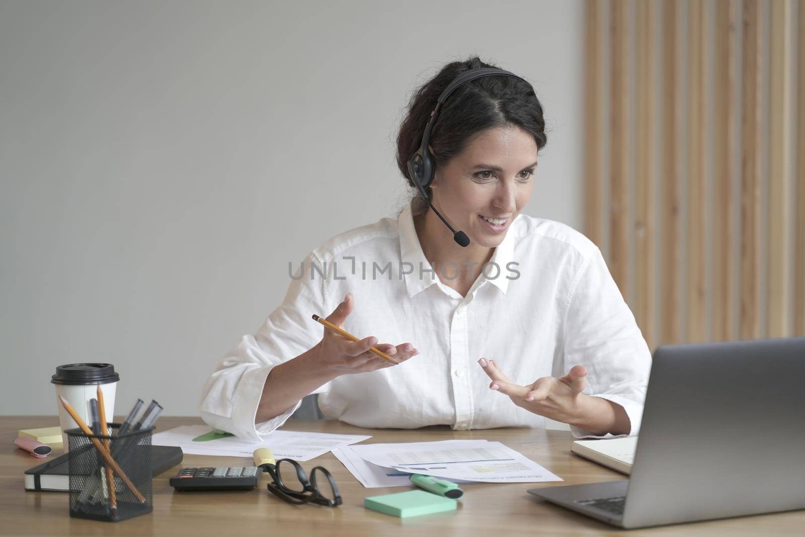 Smiling woman online tutor sitting at desk in headset and talking by video call on laptop computer by vkstock