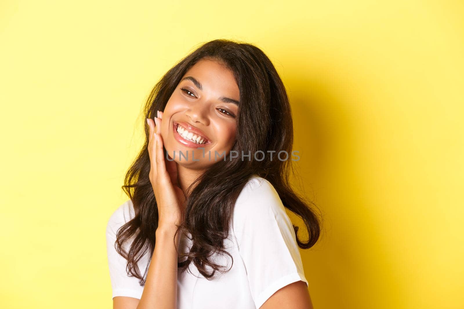 Image of gorgeous african-american woman touching her face, smiling pleased and looking left at copy space, standing over yellow background.