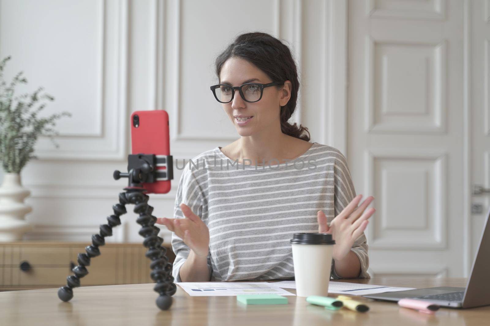 Confident Italian woman economist counselor recording video stream online consultation for group and individuals, tutor lady using phone on tripod for video chat sitting at home. Business blog concept