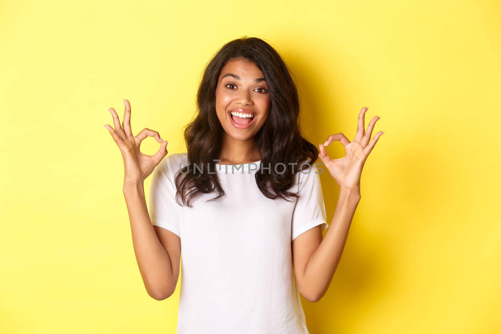Portrait of happy and pleased african-american girl, wearing white t-shirt, showing okay signs to praise excellent choice, approve and agree with something, standing over yellow background.