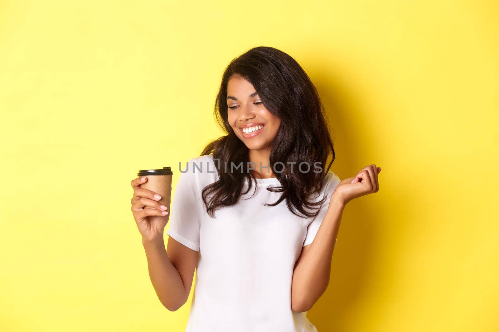 Portrait of happy and pleased african-american girl, looking at coffee and smiling, standing over yellow background.