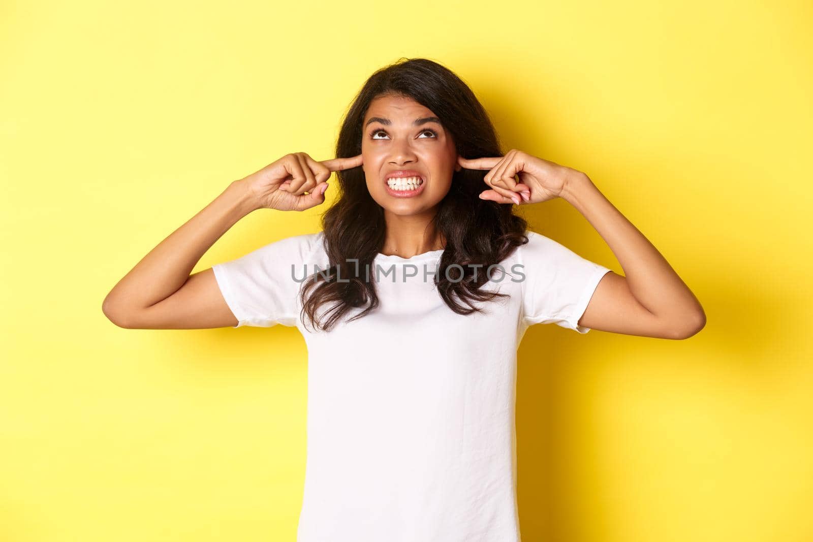 Image of annoyed and bothered african-american girl, shut ears and looking angry up, complaining at loud neighbours, standing over yellow background by Benzoix