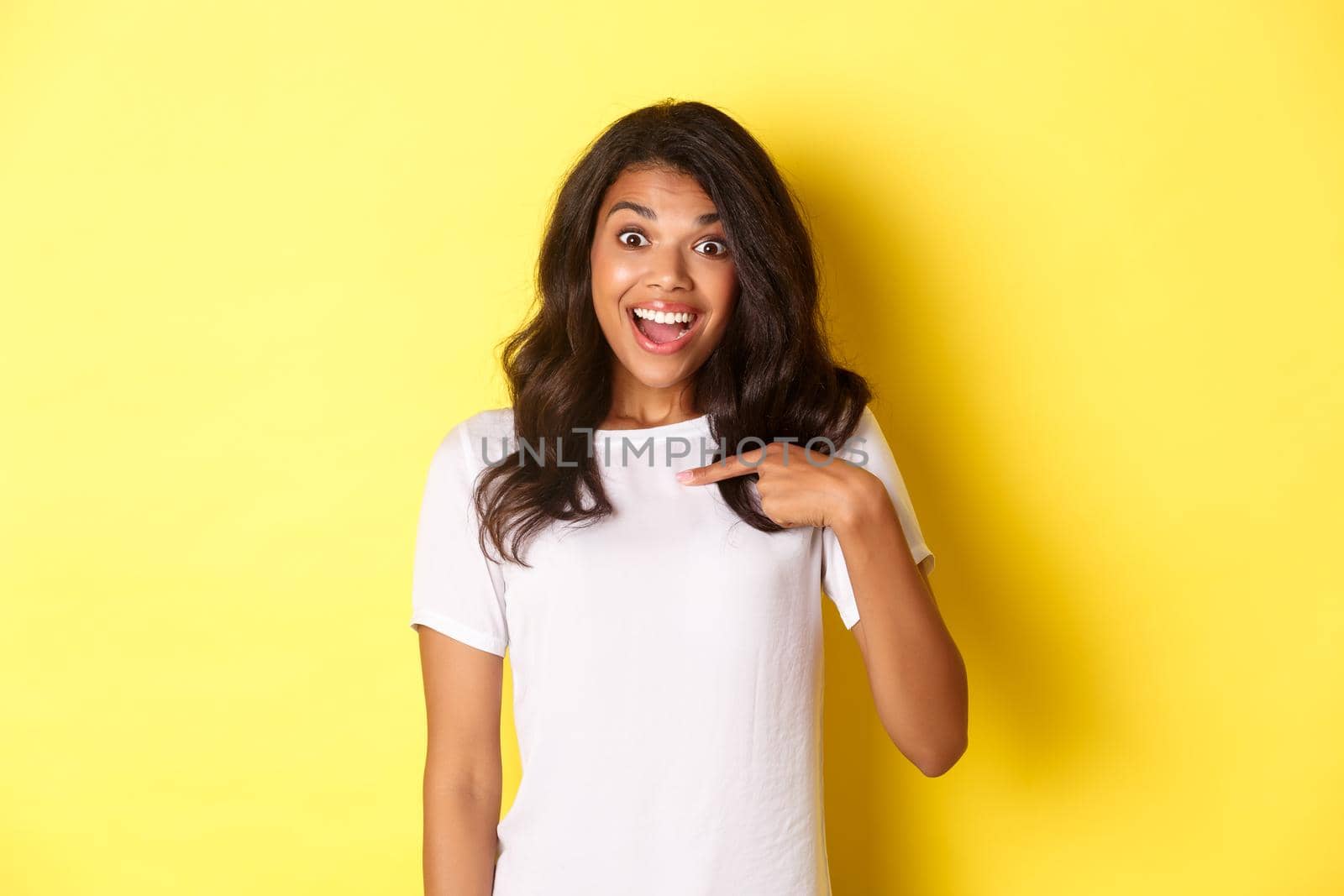Image of attractive african-american girl looking amazed and happy, smiling while pointing at herself, being chosen, standing over yellow background by Benzoix