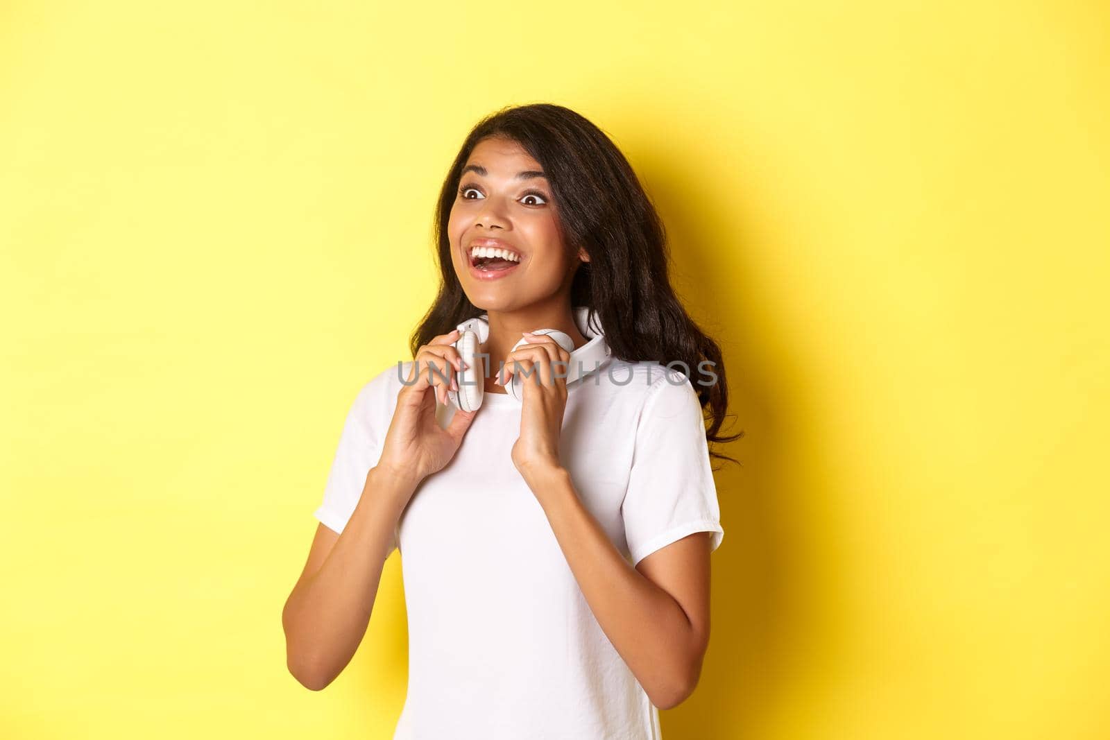 Portrait of beautiful african-american woman, holding headphones on neck and smiling, looking at upper left corner amazed, standing over yellow background.