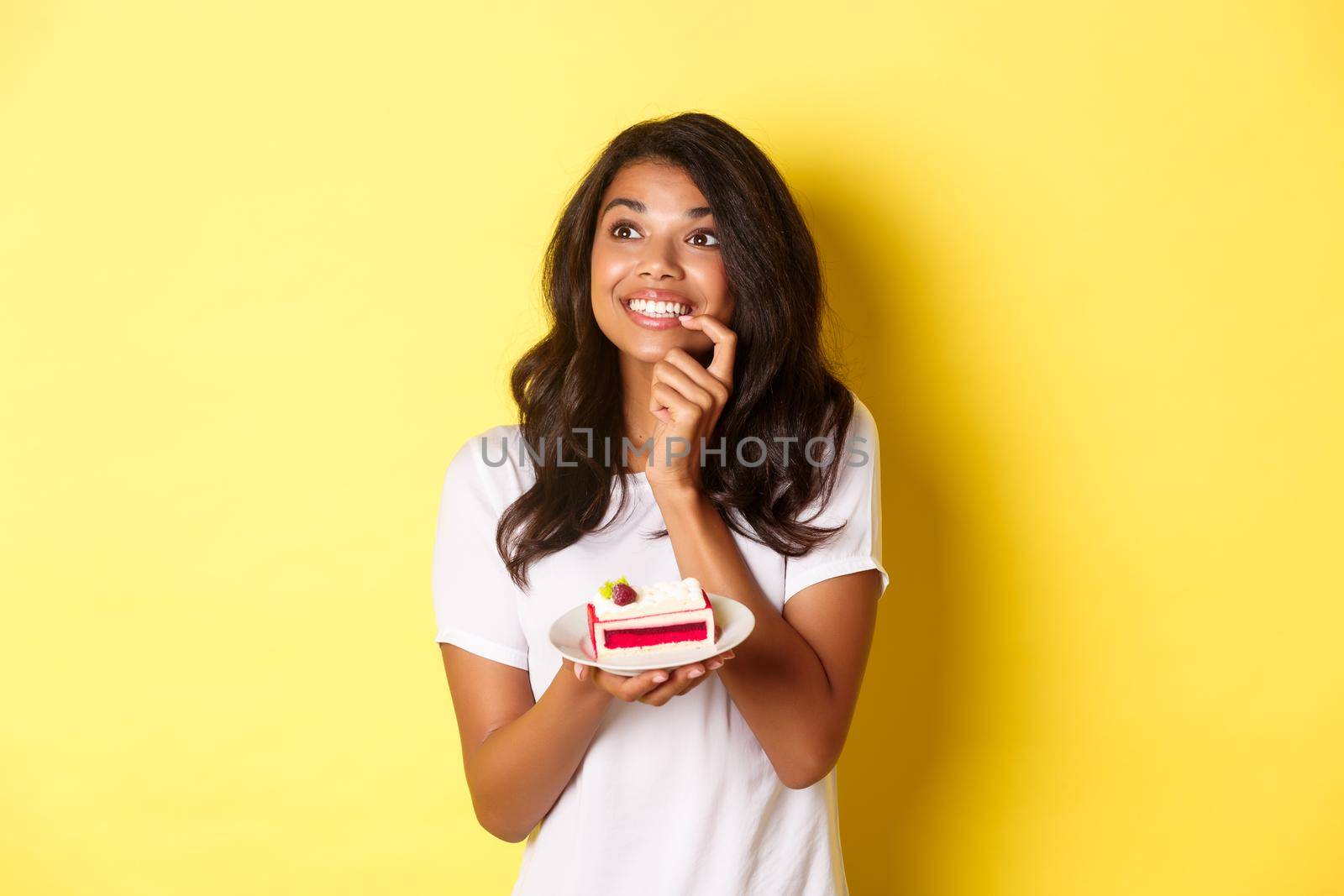 Dreamy african-american woman, smiling and looking at upper left corner thoughtful, holding piece of cake, standing over yellow background by Benzoix