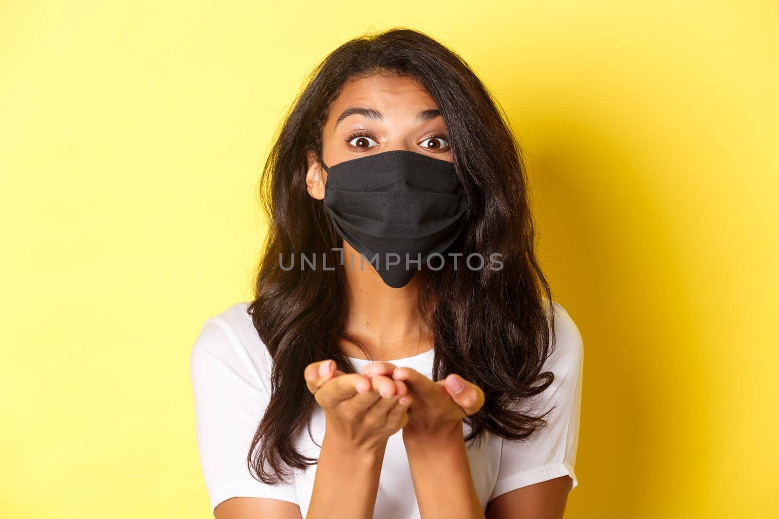 Concept of covid-19, social distancing and lifestyle. Close-up of lovely african-american girl in black face mask, giving something, holding in hands, standing over yellow background.