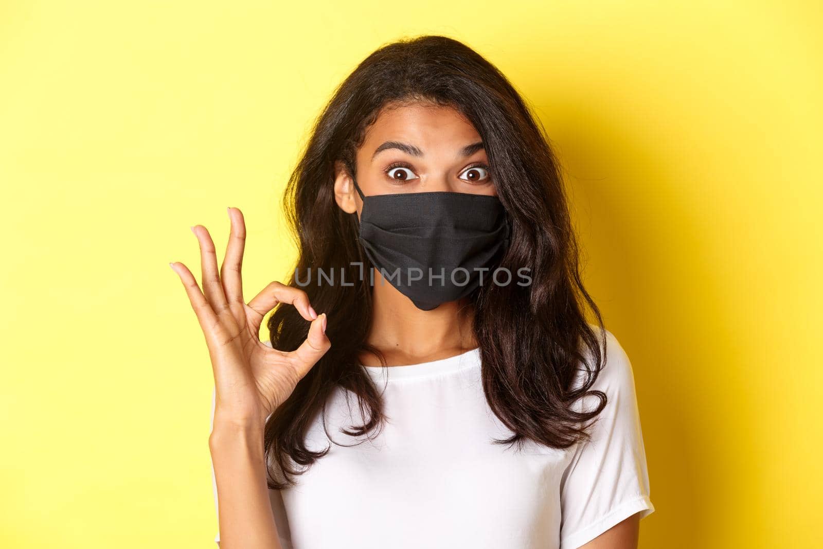 Concept of coronavirus, pandemic and lifestyle. Close-up of amazed and satisfied african-american girl, wearing black face mask, showing okay sign, like something good, yellow background.