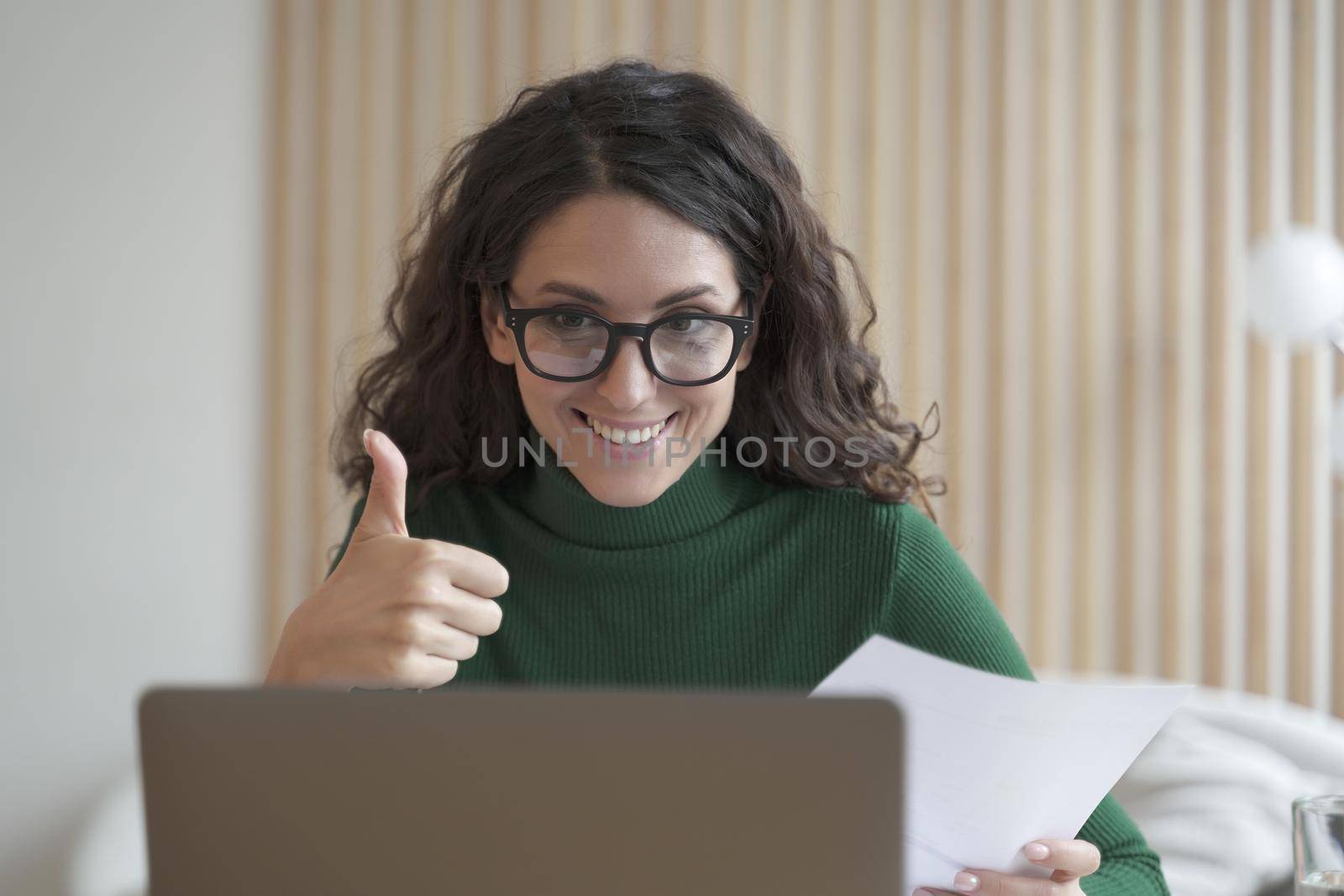 Happy young Italian female employee talks online on laptop with colleagues and showing thumb up by vkstock