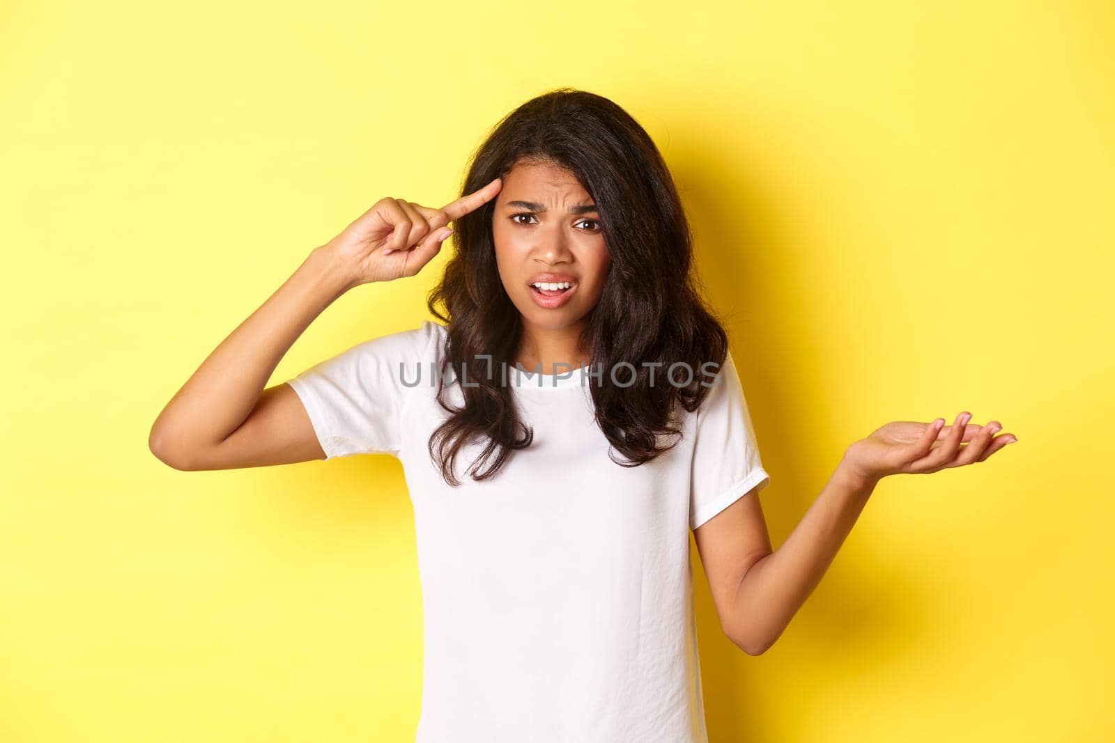 Portrait of angry and confused african-american girl, scolding someone for being stupid, pointing at head and shrugging, standing over yellow background.