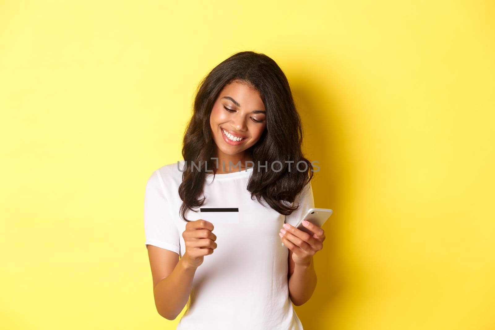 Young attractive woman shopping online, using mobile phone with credit card and smiling, standing over yellow background by Benzoix