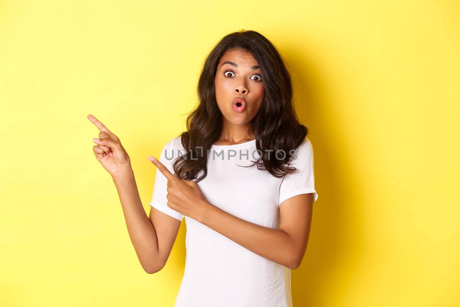 Portrait of surprised african american girl in white t-shirt, looking amazed and pointing fingers at upper left corner, showing your logo or promo banner over yellow background by Benzoix