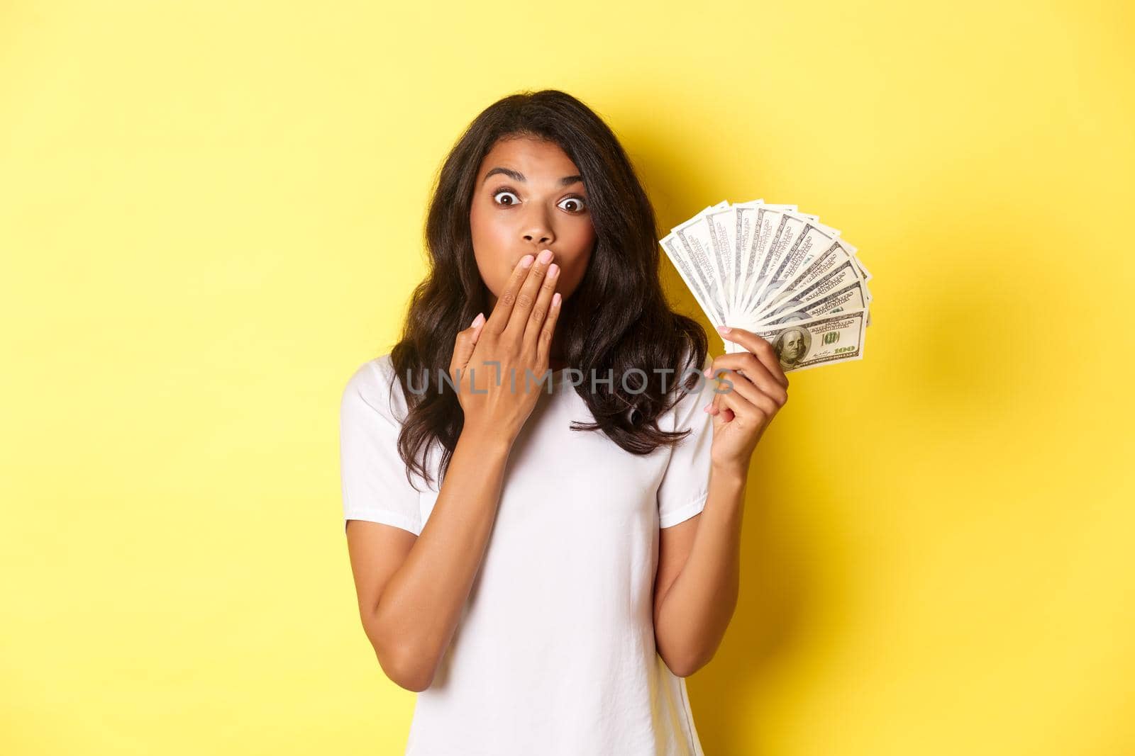 Image of surprised african-american girl winning money, gasping amazed, standing over yellow background.