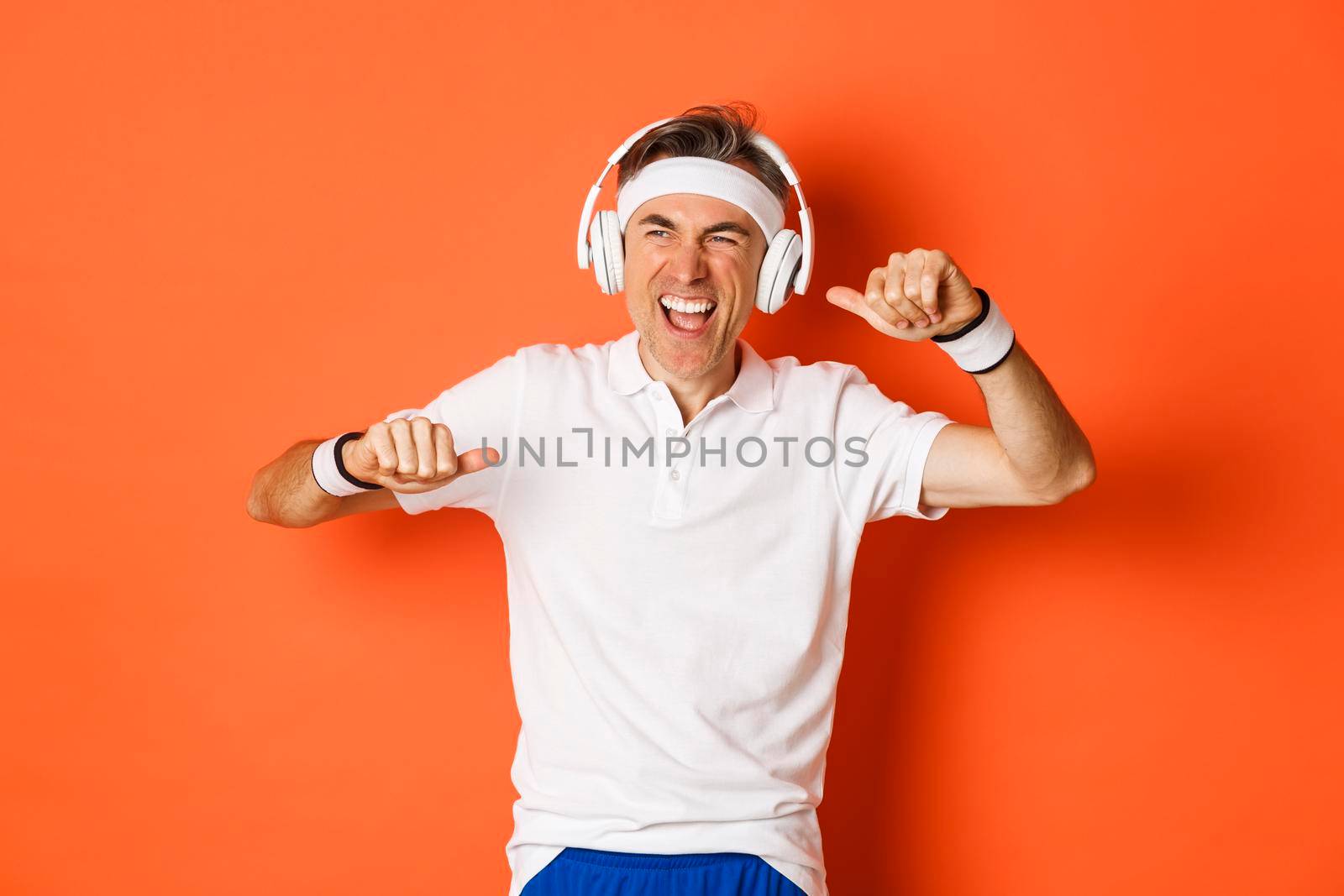 Portrait of handsome middle-aged male athlete, listening music in headphones during fitness training, dancing over orange background by Benzoix