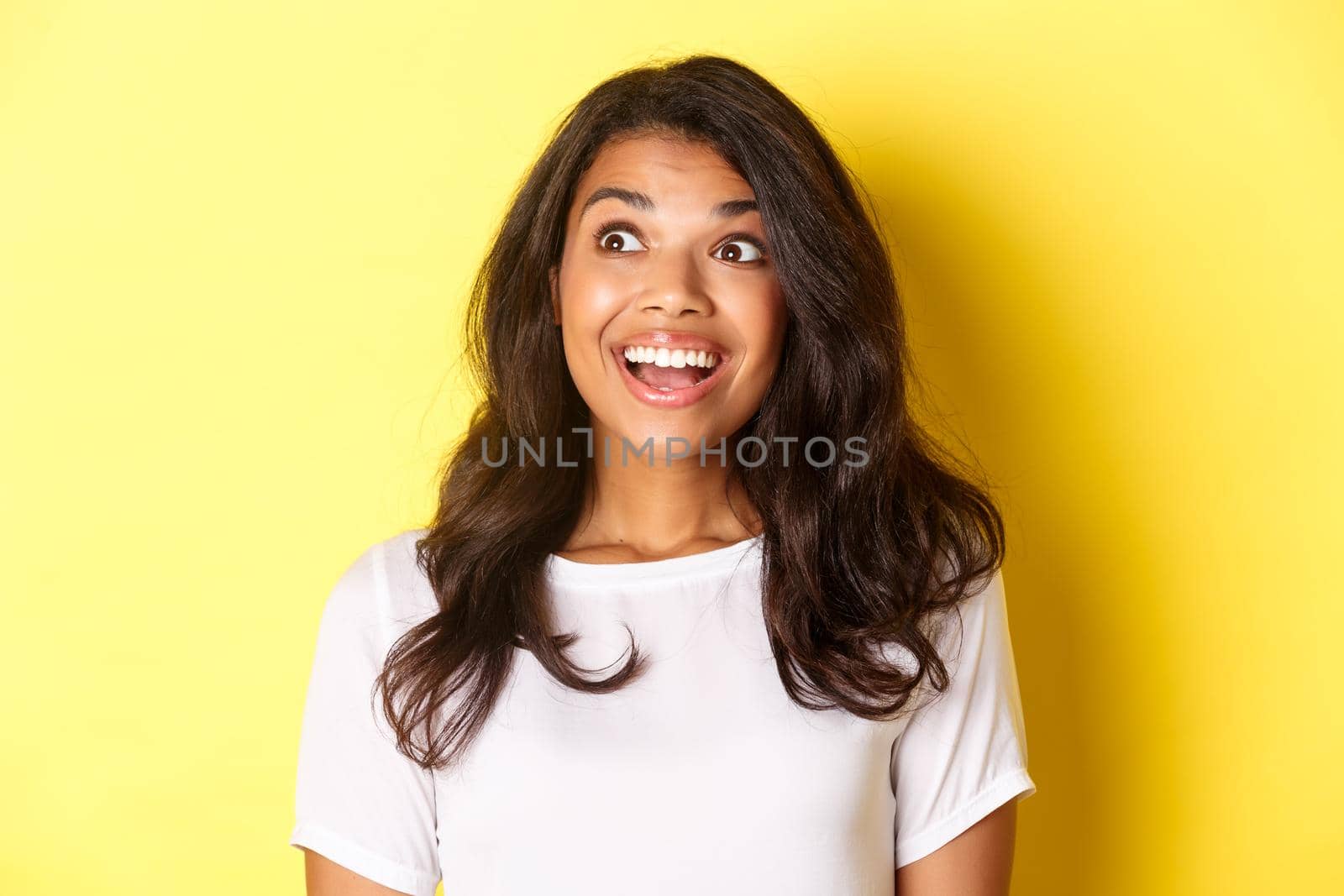 Image of excited and hopeful african-american girl in white t-shirt, smiling and looking amazed at upper left corner advertisement, standing over yellow background.