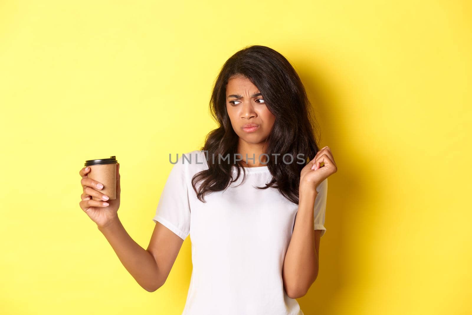 Portrait of african-american girl looking disappointed at cup, dont like bad coffee, standing over yellow background by Benzoix
