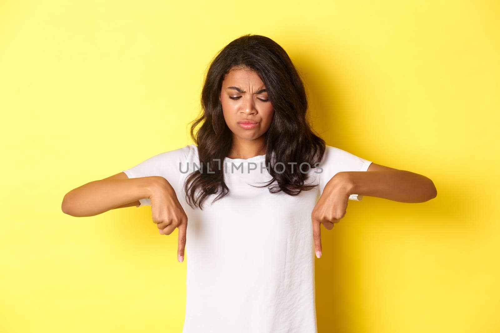 Portrait of sad and disappointed african-american girl, complaining about something bad, looking and pointing fingers down with displeased face, standing over yellow background by Benzoix