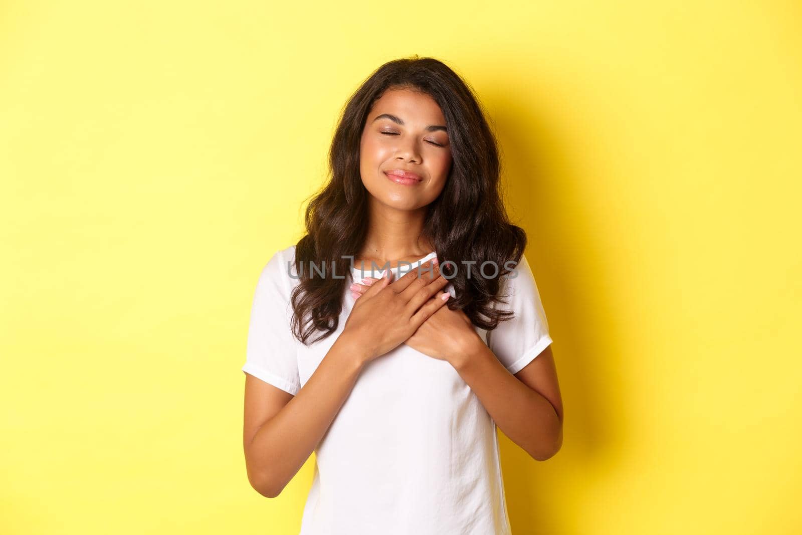 Portrait of romantic african-american woman in white t-shirt, close eyes and hold hands on heart, feeling nostalgic, thinking about someone or having beautiful memory, yellow background.
