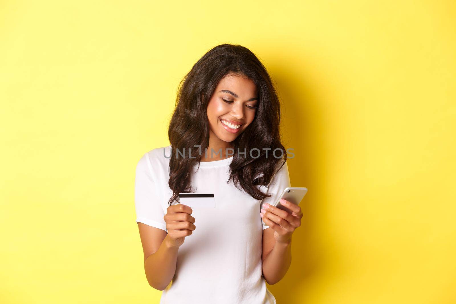 Portrait of good-looking african american woman, shopping online with smartphone and credit card, standing over yellow background.