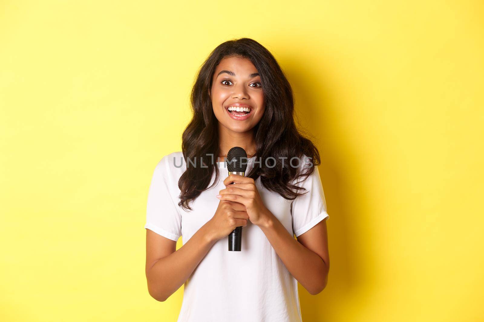 Portrait of happy african-american girl, looking amused while giving speech, holding microphone and smiling at camera, standing over yellow background.