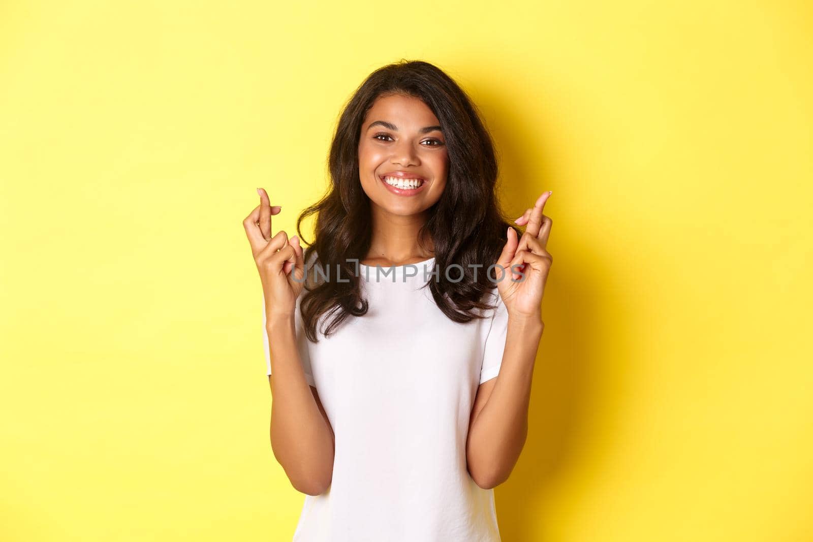 Portrait of optimistic, smiling african-american girl, crossing fingers for good luck and making wish, standing over yellow background by Benzoix