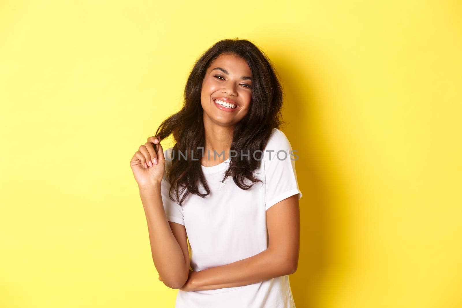 Portrait of attractive and confident african-american woman, wearing white t-shirt, smiling happy and playing with hair, standing over yellow background.