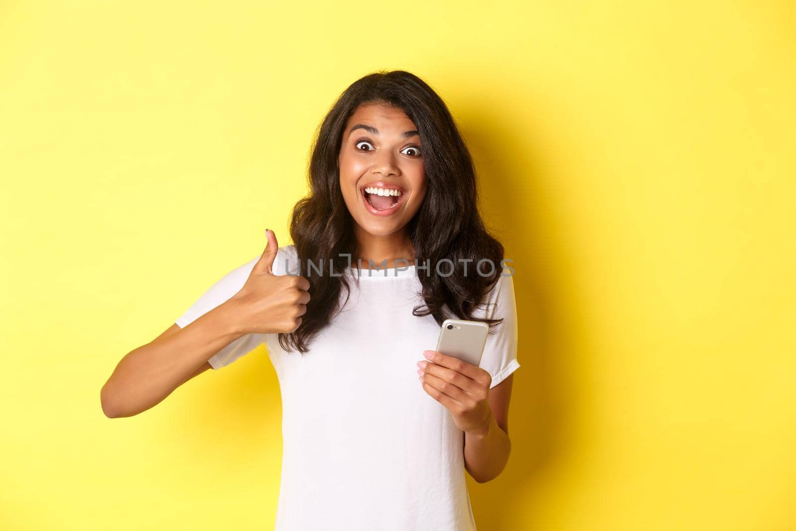 Portrait of amazed african-american girl, showing thumbs-up and using mobile phone, praising cool app, standing over yellow background by Benzoix