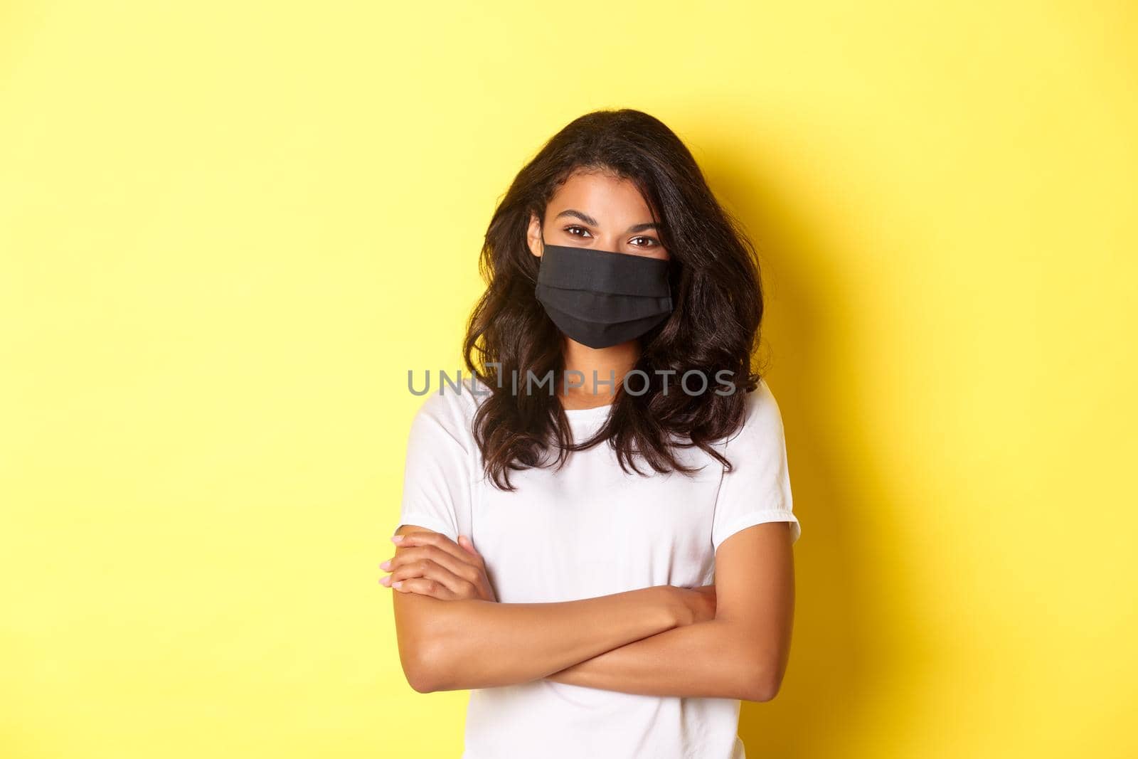 Concept of coronavirus, pandemic and lifestyle. Portrait of young african-american woman in black face mask, smiling and looking confident with hands crossed on chest, yellow background by Benzoix