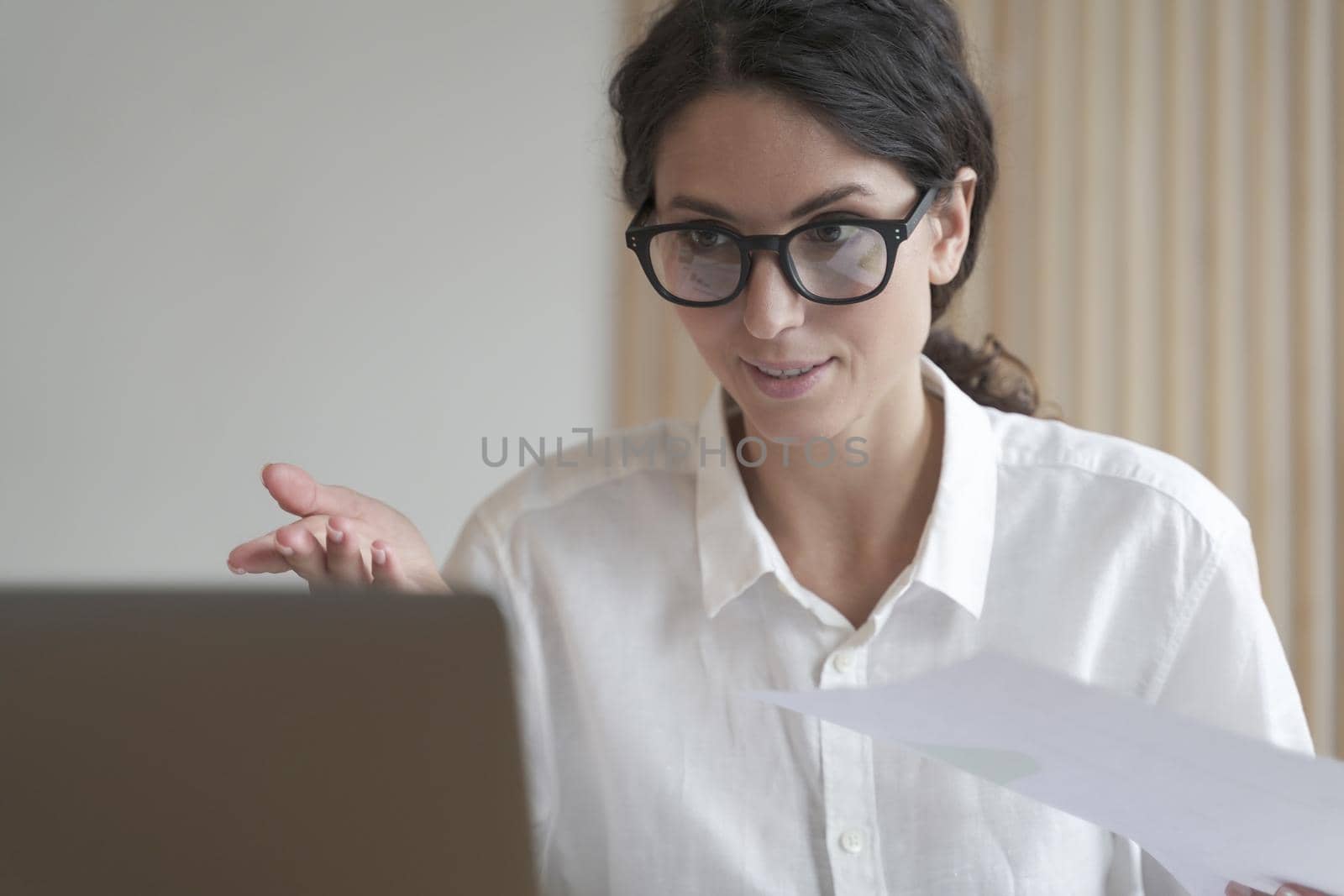 Confident woman in glasses sitting at desk, works on computer at home. Concentrated business lady looks at laptop screen gestures to communicate with client via video chat, operates remotely online