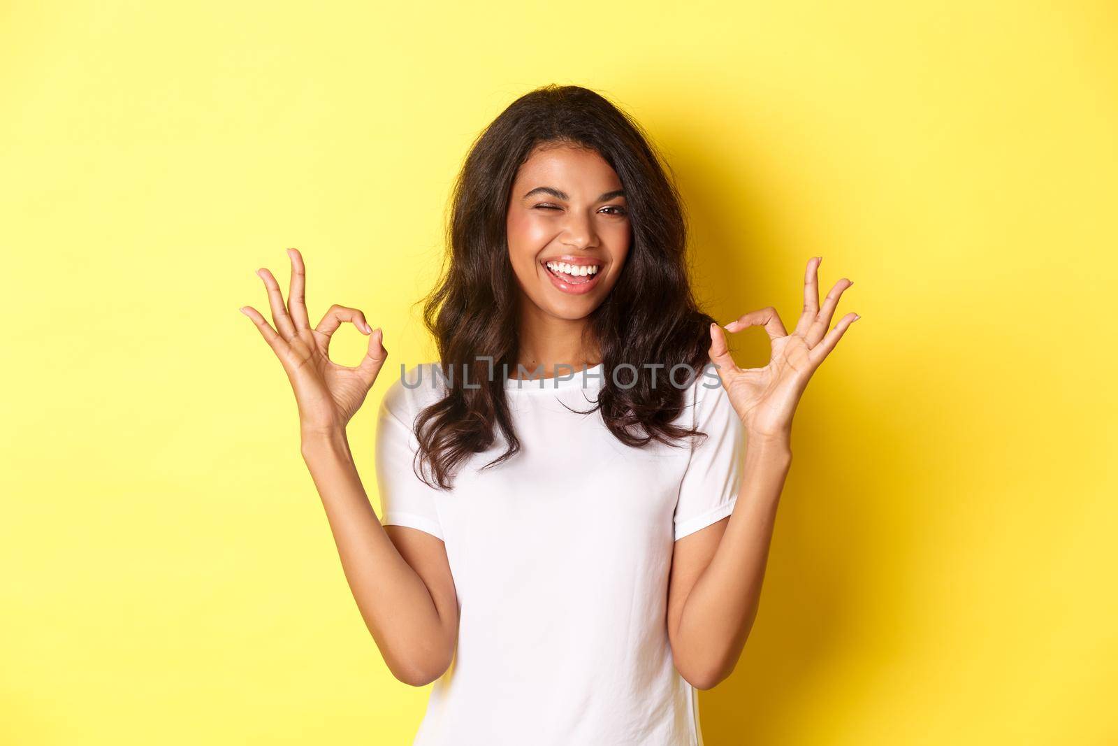Portrait of confident, attractive african-american girl, showing okay signs and winking, guarantee quality, approve and agree something good, standing over yellow background by Benzoix