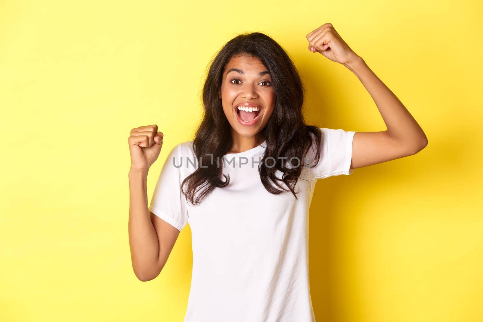 Portrait of cheerful african-american girl, winning and celebrating victory, raising hands up to rejoice, standing over yellow background.