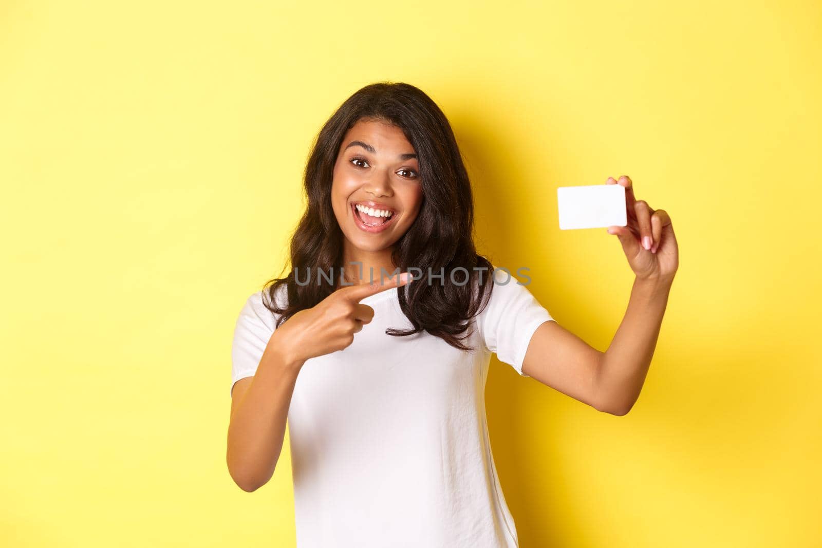 Image of beautiful african-american woman in white t-shirt, pointing at credit card and smiling, recommending bank, standing over yellow background by Benzoix
