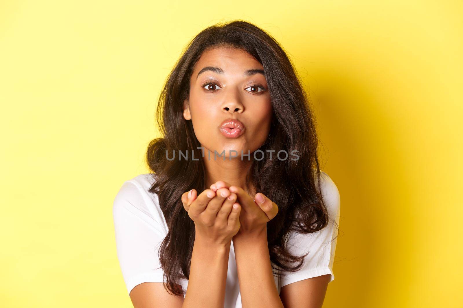 Close-up of beautiful and romantic african-american woman, pucker lips and sending air kiss, standing against yellow background by Benzoix