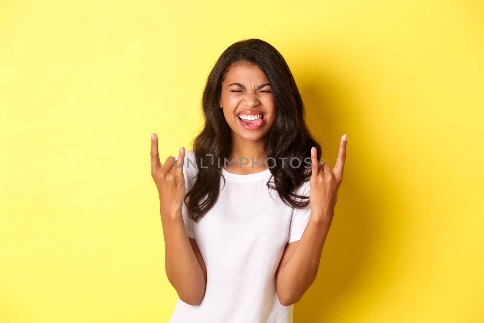 Image of excited and carefree african-american girl, enjoying something cool, making rock-on signs and smiling, standing over yellow background.