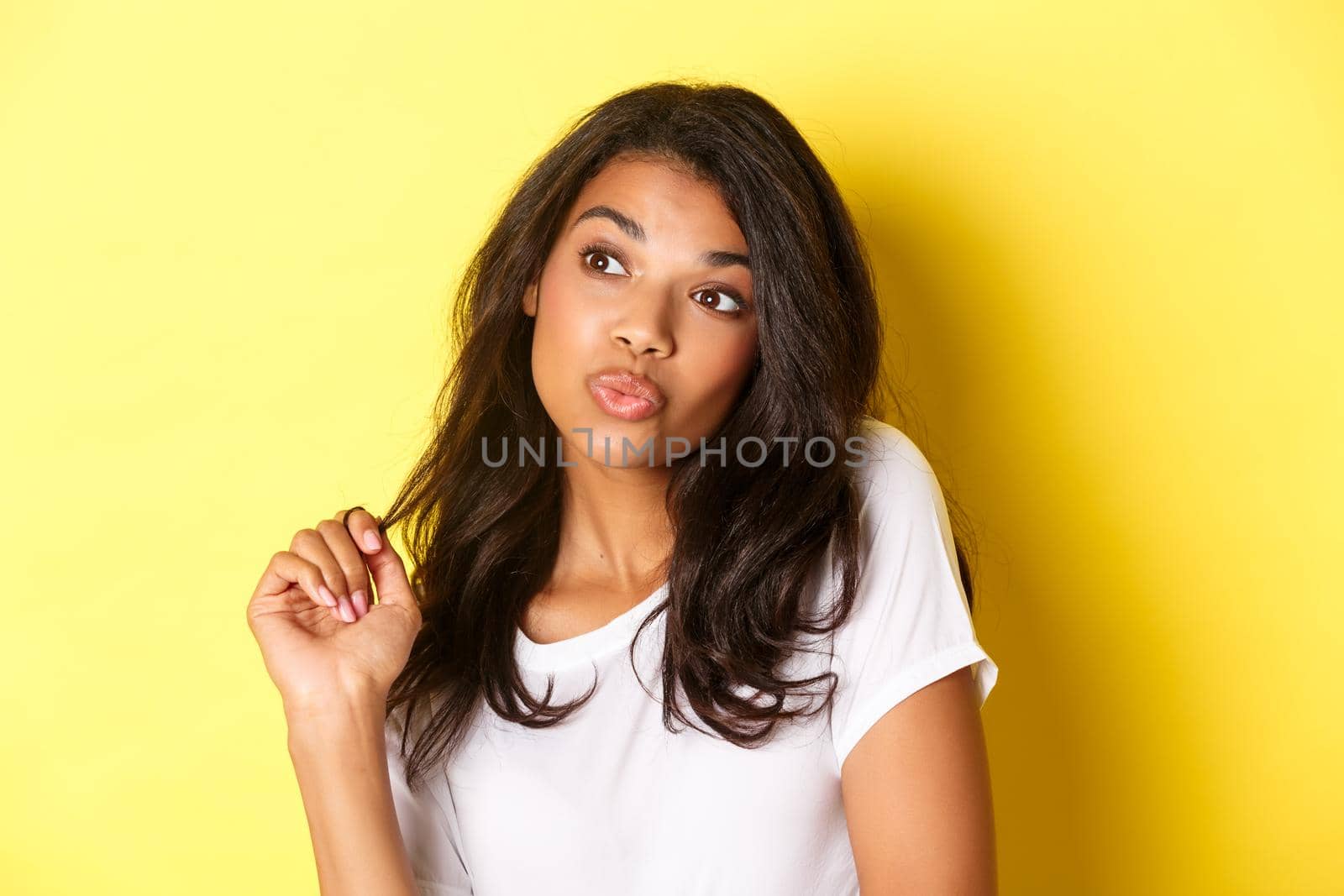 Image of silly and cute african-american girl, pouting and looking left, standing over yellow background.