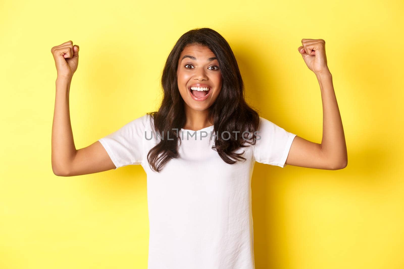 Image of happy african-american girl achieve goal and celebrating victory, raising hands up and smiling pleased, looking satisfied, standing over yellow background.